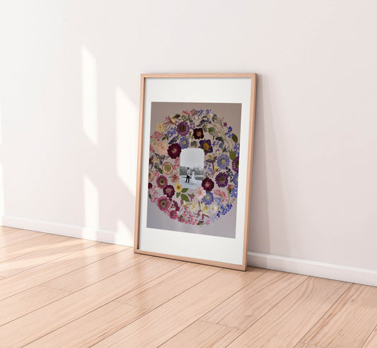A framed art print leaning against a white wall on a light wood floor. The print features a circular floral wreath surrounding a black-and-white photograph of a person, and it is housed in a light natural wood frame with a white mat.