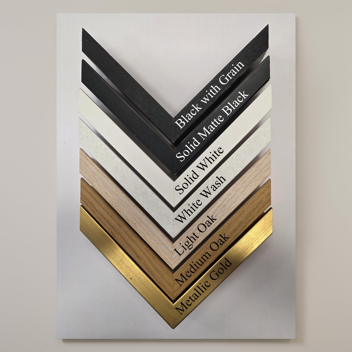 A vertical wall-mounted sample display showing chevron-shaped wood and metal frame finishes labeled Black with Grain, Solid Matte Black, Solid White, White Wash, Light Oak, Medium Oak, and Metallic Gold, on a light neutral background.