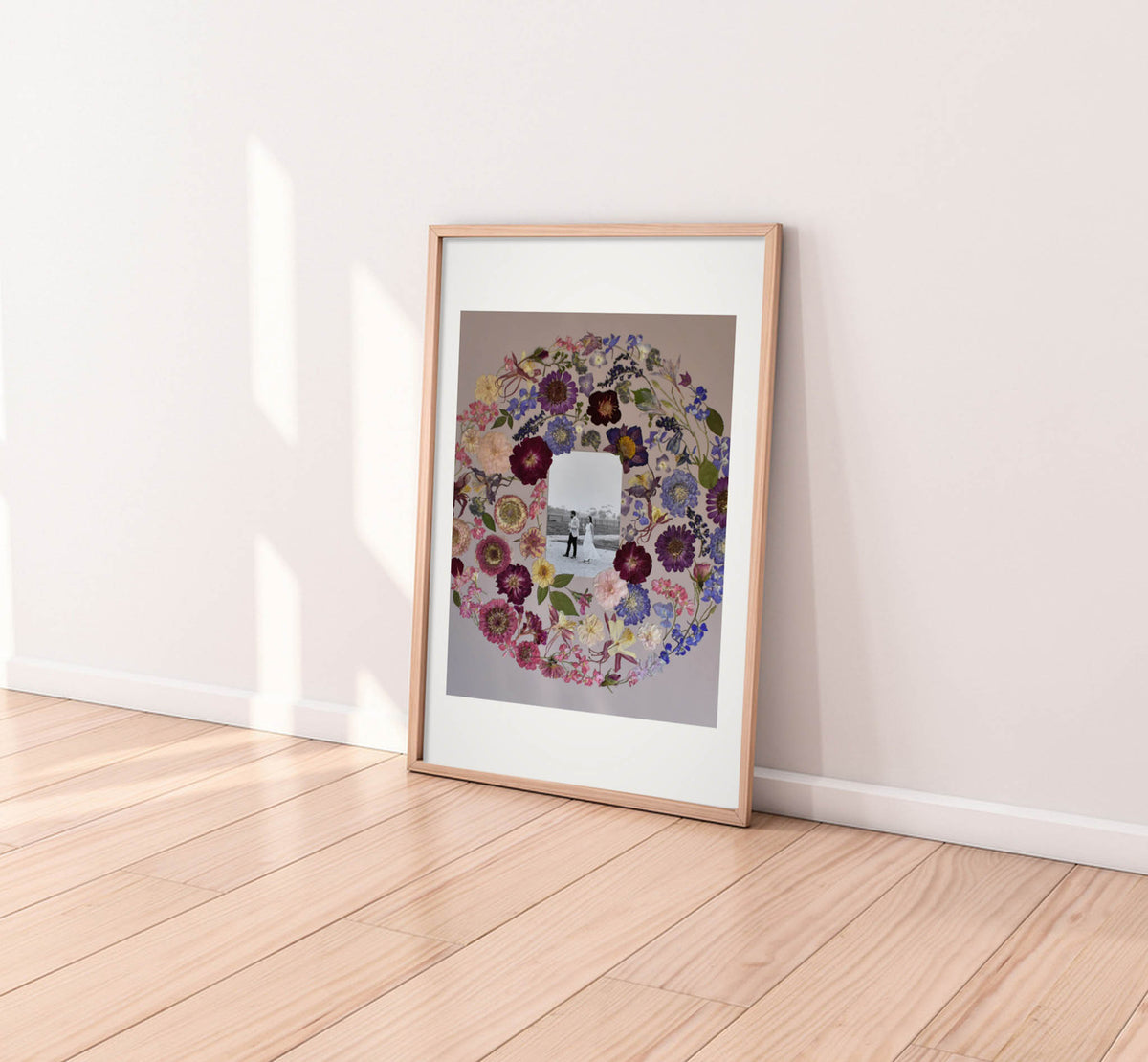 A framed art print leaning against a white wall on a light wood floor. The print features a circular floral wreath surrounding a black-and-white photograph of a person, and it is housed in a light natural wood frame with a white mat.