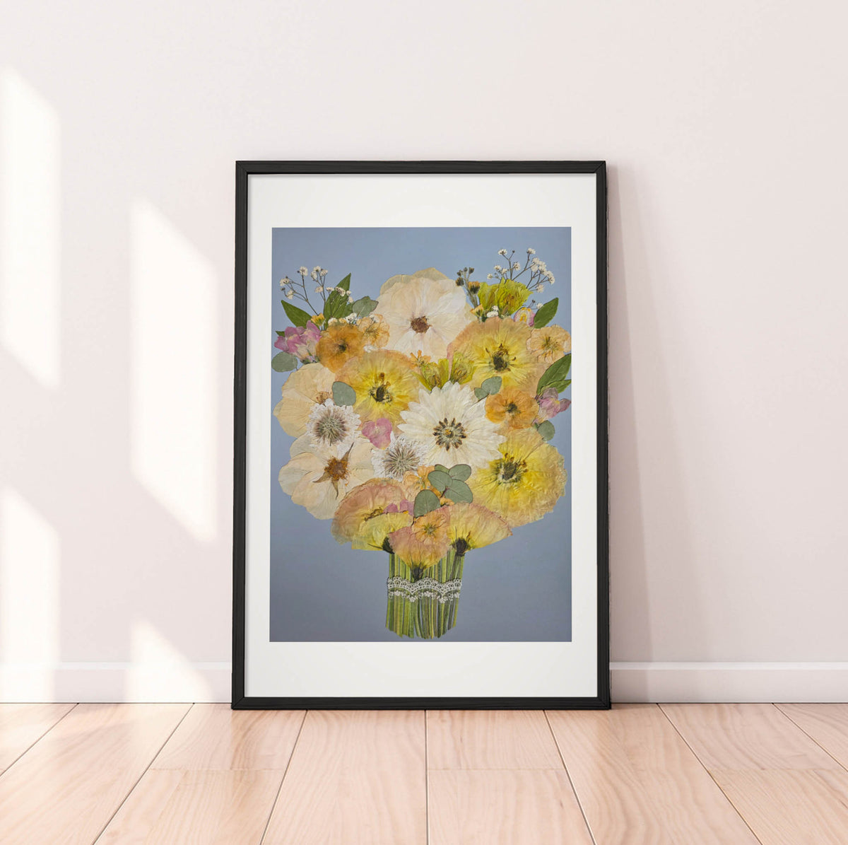 A framed floral art print leaning against a white wall on a light wood floor. The artwork shows a bouquet of yellow, white, and pink flowers with green leaves on a soft blue-gray background, enclosed in a black frame with a white mat.