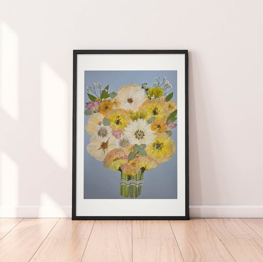 A framed floral art print leaning against a white wall on a light wood floor. The artwork shows a bouquet of yellow, white, and pink flowers with green leaves on a soft blue-gray background, enclosed in a black frame with a white mat.
